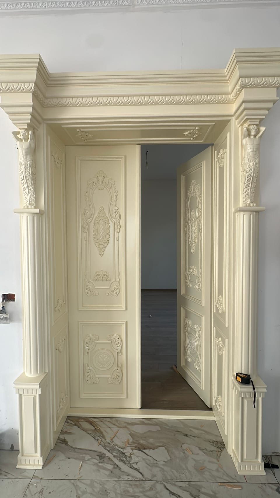 Ușă dublă de culoare fildeș, întredeschisă, cu panouri cu ornamente în relief și coloane cu figuri în relief pe ancadrament.