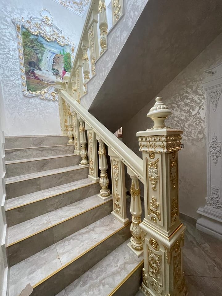 Scară cu balustradă crem, baluștri cu ornamente aurii, mână curentă și stâlp de pornire decorat, lângă o pictură murală.