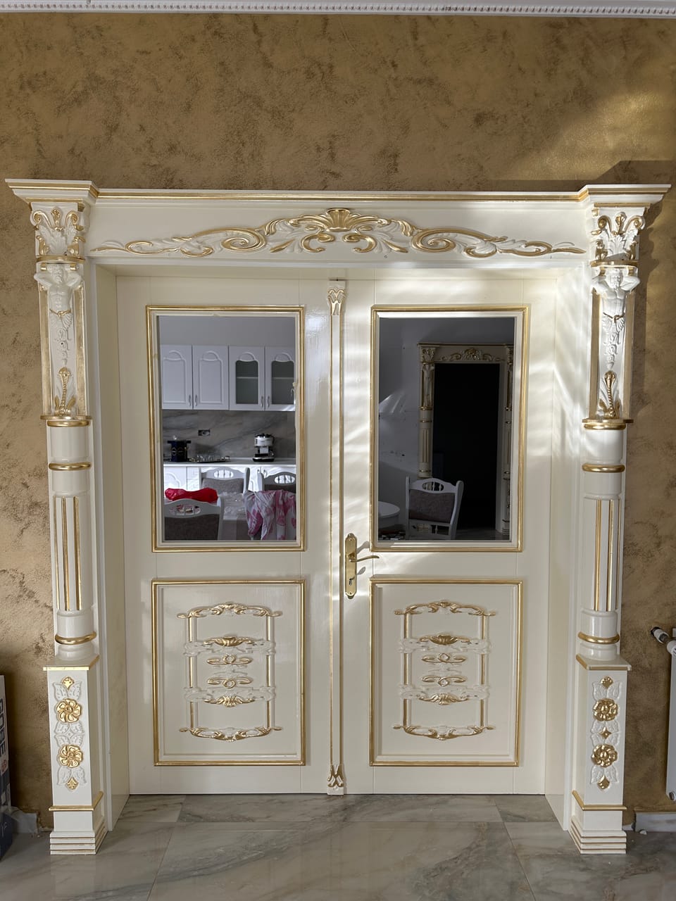Ușă dublă albă cu geamuri, accente aurii, ornamente în relief și coloane cu figuri în relief pe ancadrament.