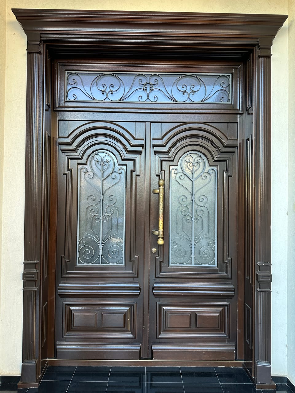 Ușă dublă maro închis cu arcade, geamuri cu feronerie decorativă, supralumină și mâner auriu vertical.