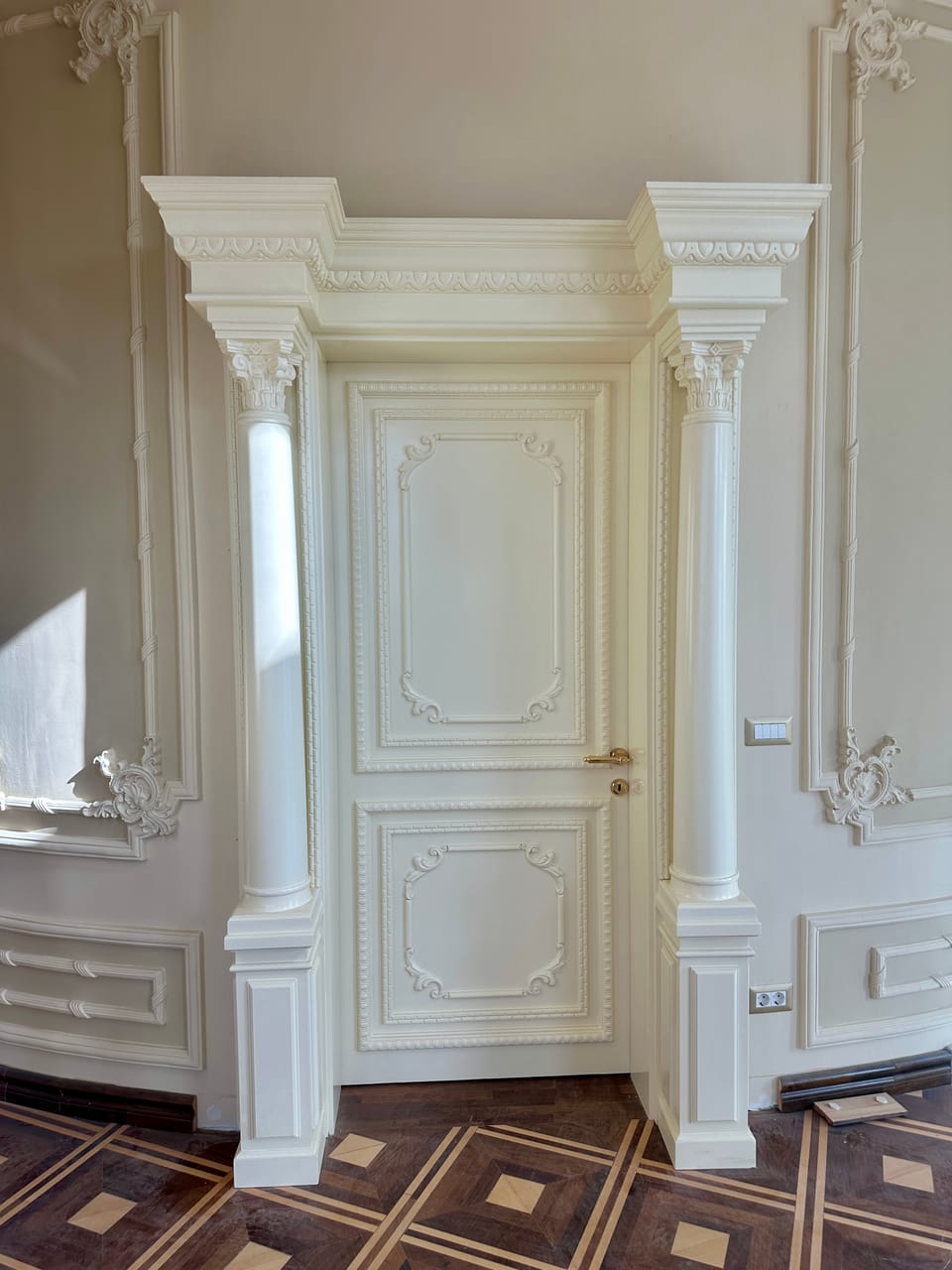 Ușă albă cu panouri profilate și mâner auriu, încadrată de coloane și o cornișă cu ornamente în relief.