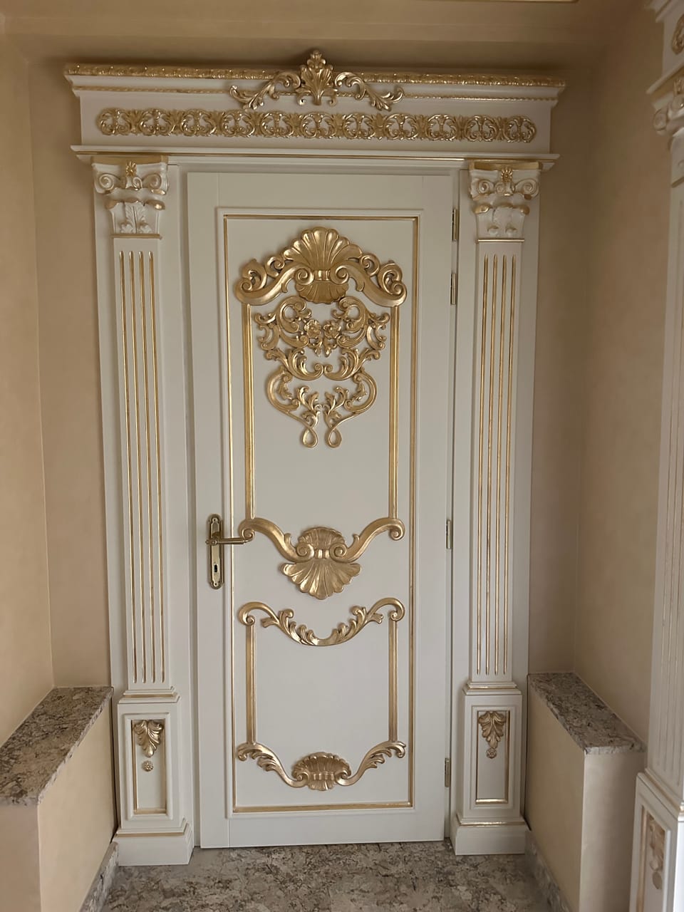 Ușă albă cu ornamente aurii în relief, mâner auriu și ancadrament cu pilaștri canelați și cornișă decorată.