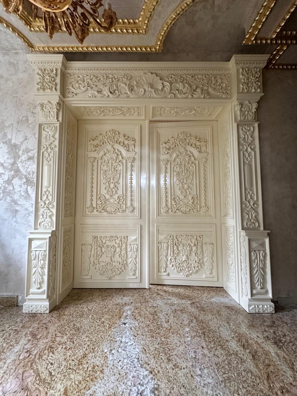 Ușă dublă crem, întredeschisă, cu panouri și ancadrament acoperite de ornamente bogate în relief.