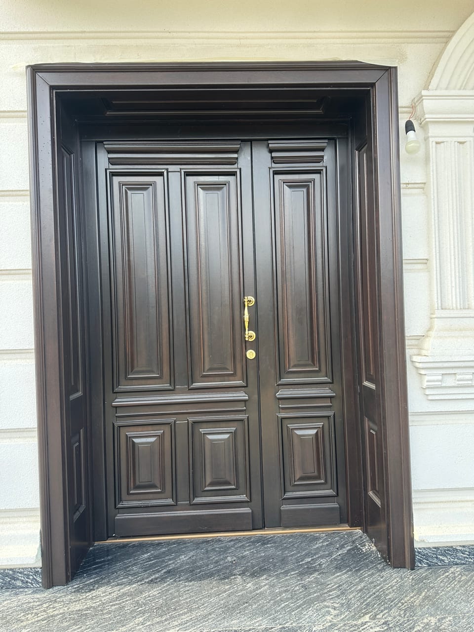 Ușă dublă de intrare maro închis, cu panouri profilate, ancadrament adânc și mâner auriu, pe o fațadă deschisă la culoare.