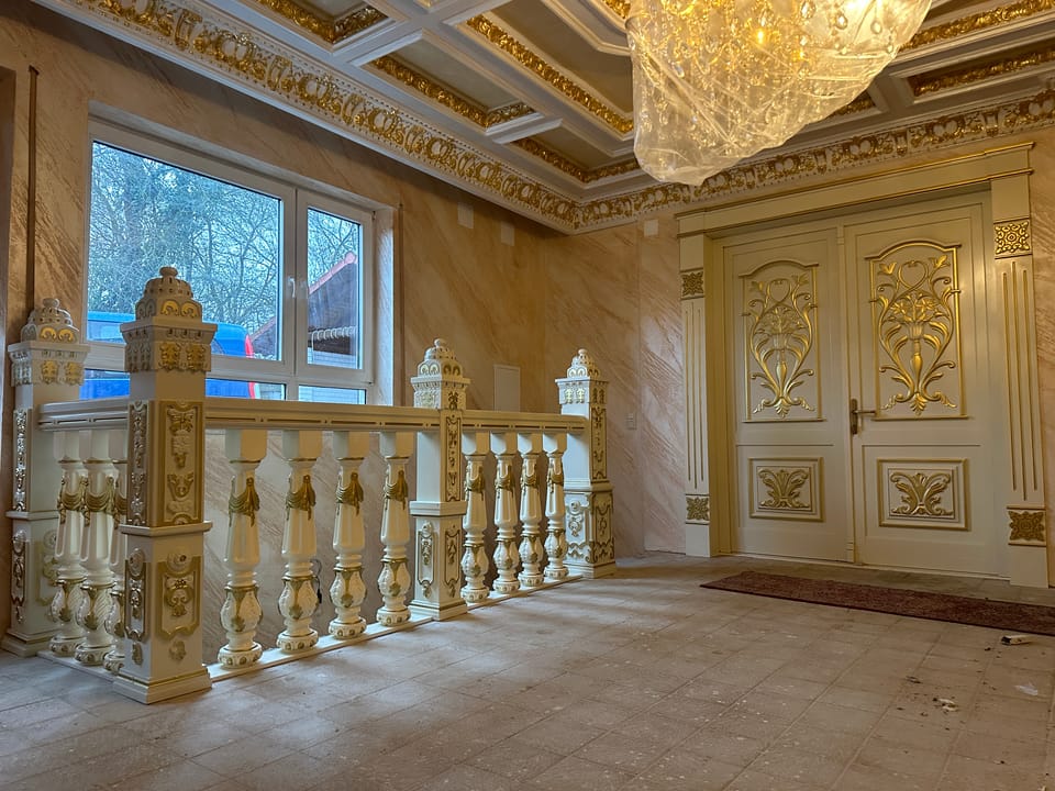 Balustradă crem cu baluștri și stâlpi cu ornamente aurii în relief, în fața unei uși duble cu ornamente florale aurii.