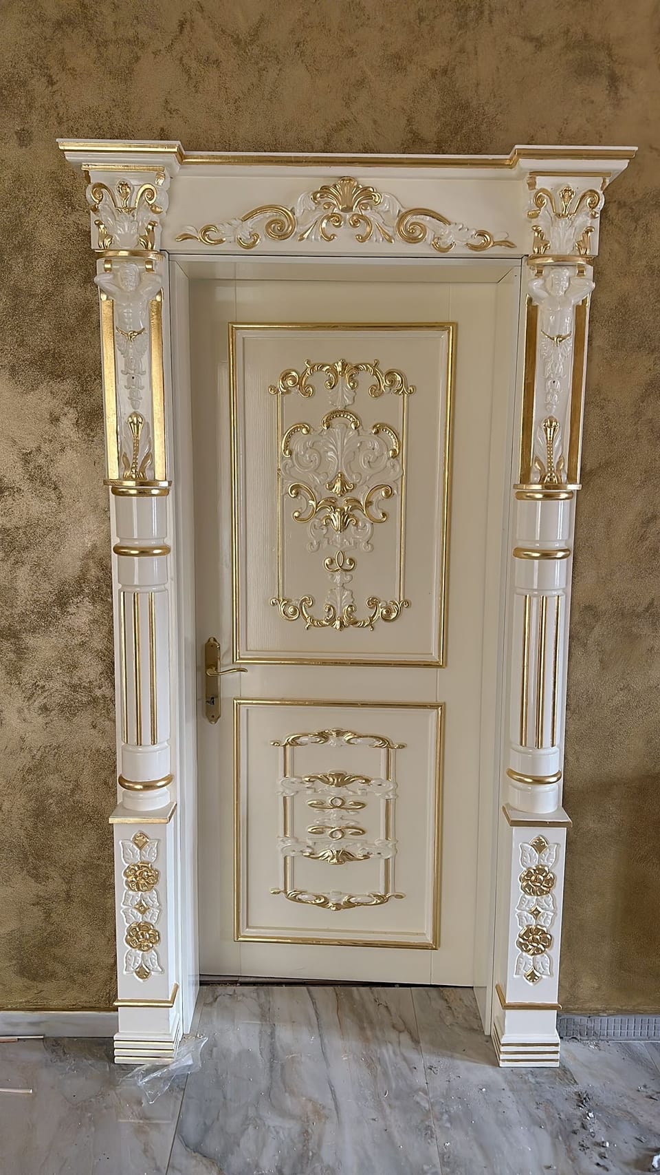 Ușă crem cu ornamente aurii și albe în relief, încadrată de pilaștri cu figuri decorative și o cornișă.