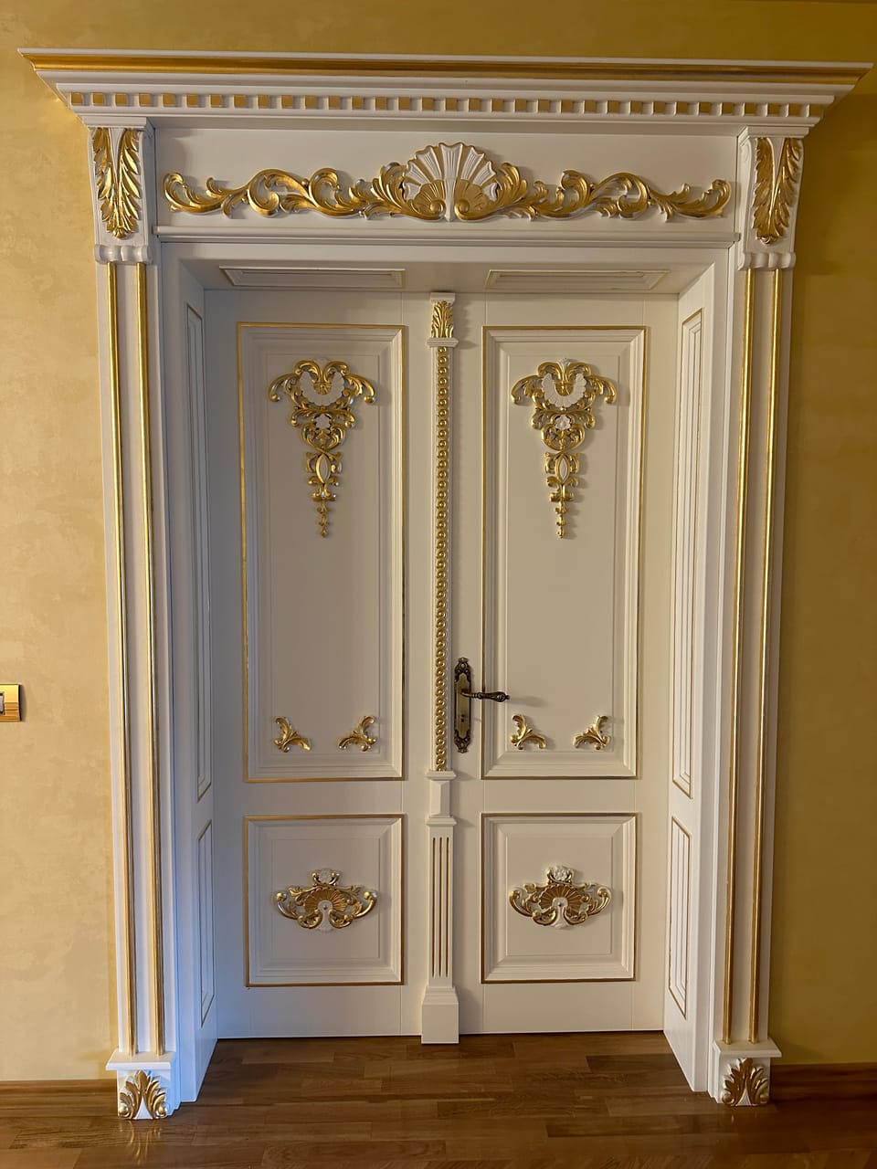 Ușă dublă albă cu panouri profilate, ornamente aurii în relief și ancadrament cu cornișă decorată.