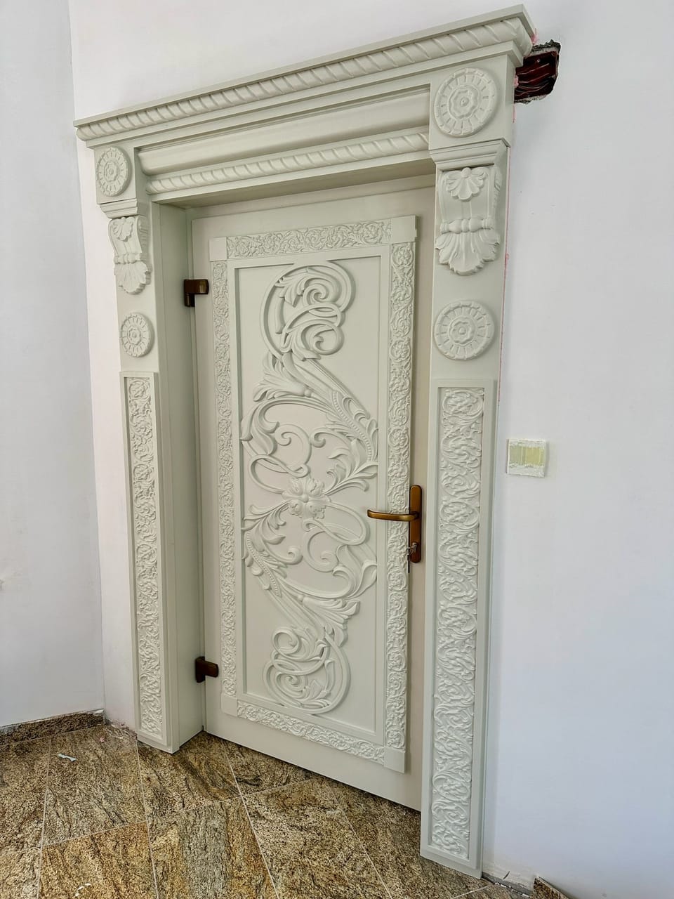 Ușă albă cu panou cu volute în relief, într-un ancadrament cu rozete, console și benzi cu ornament în relief.