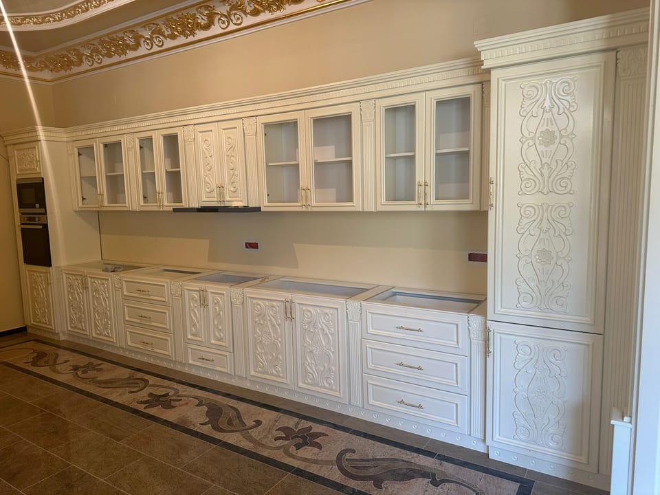 Mobilier de bucătărie crem cu fronturi cu ornamente în relief, vitrine cu geamuri, sertare și coloană înaltă decorată.