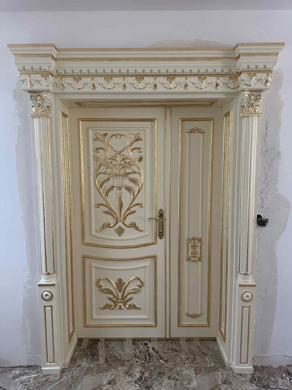 Ușă crem cu două canaturi inegale, ornamente aurii în relief și ancadrament cu cornișă decorată.