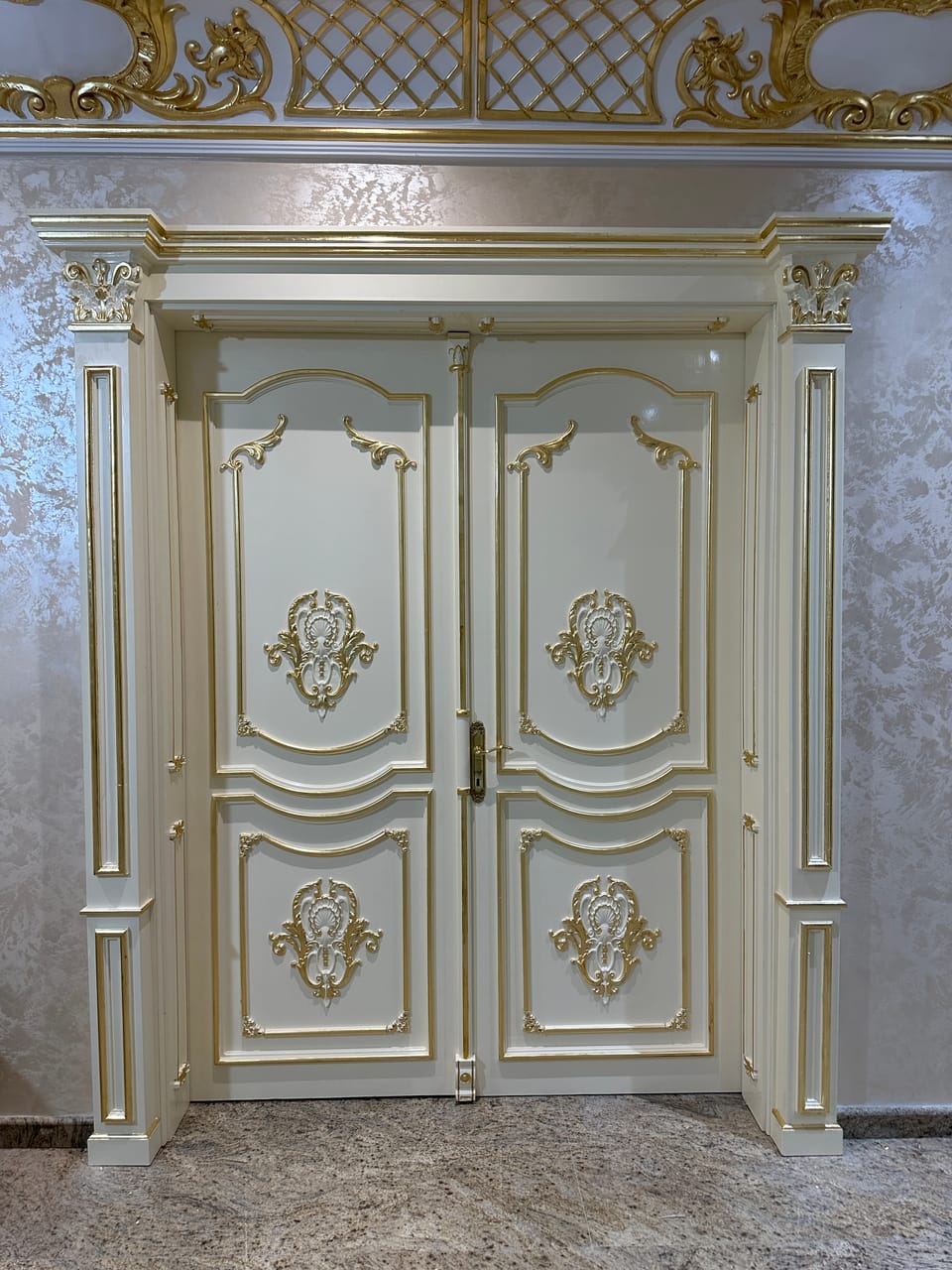 Ușă dublă albă cu ornamente aurii în relief, panouri profilate, mâner auriu și ancadrament cu pilaștri, într-un interior.