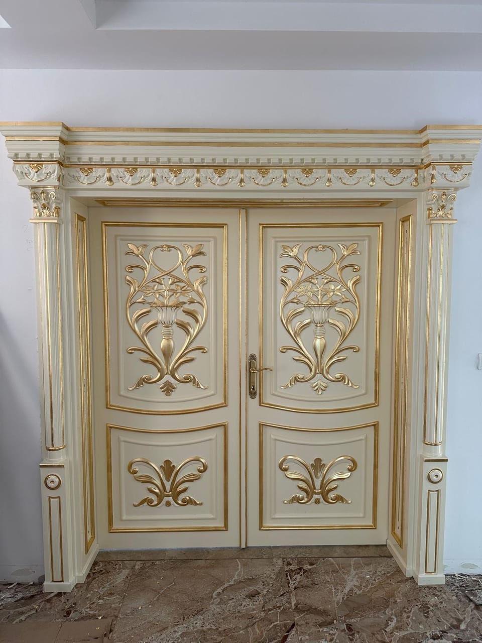 Ușă dublă crem cu ornamente florale aurii în relief, mâner auriu și ancadrament cu cornișă bogat decorată.