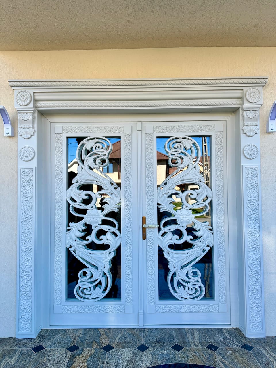 Ușă dublă albă de intrare, cu geamuri mari acoperite de feronerie decorativă în volute și ancadrament cu ornamente în relief.