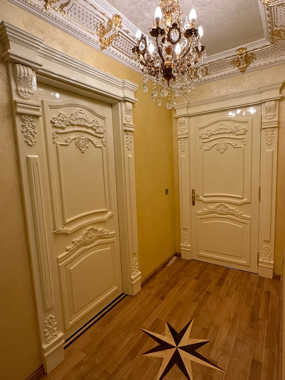 Două uși crem cu panouri profilate, ornamente în relief și ancadramente cu console, într-un hol cu candelabru.