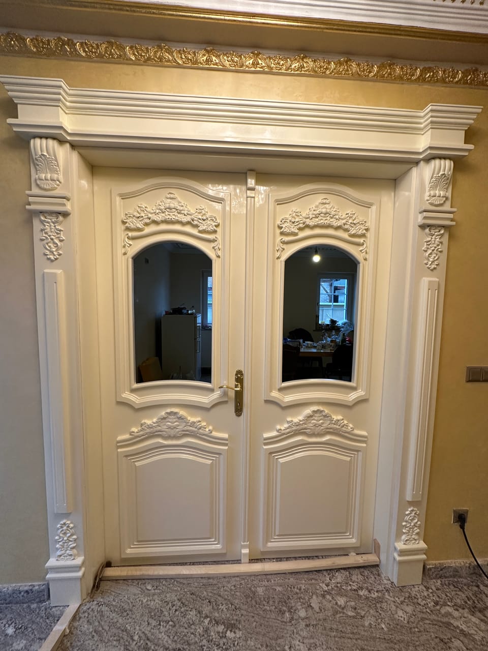 Ușă dublă crem cu geamuri arcuite, ornamente în relief, panouri profilate și mâner auriu, într-un ancadrament cu console.
