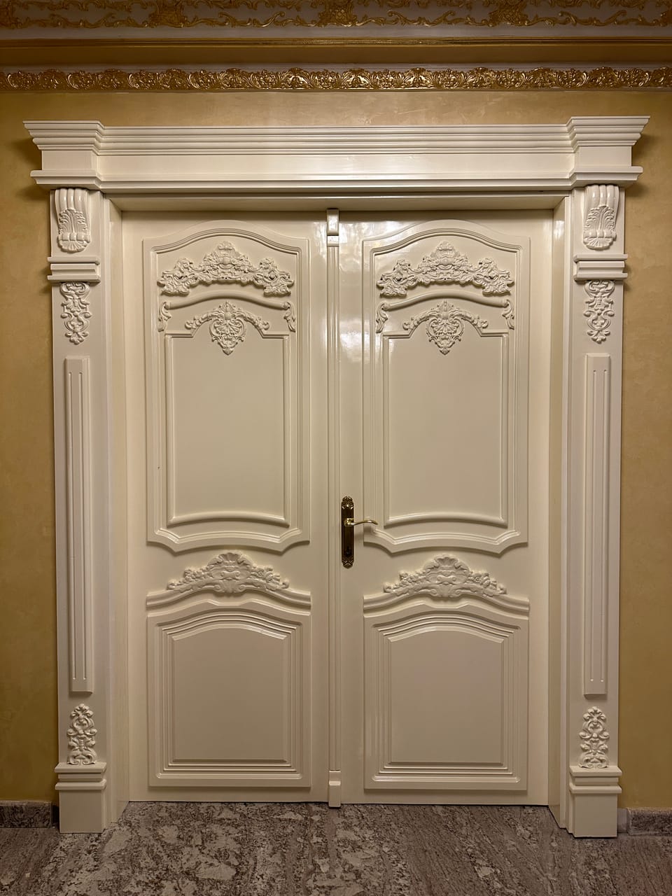 Ușă dublă crem cu panouri profilate, ornamente în relief și mâner auriu, într-un ancadrament cu console și pilaștri.